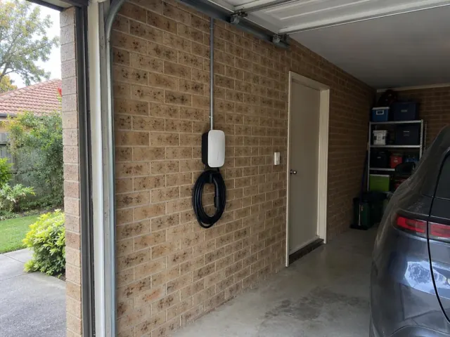 Wall-mounted EV charger on a brick garage wall with conduit and coiled cable in a carport in Box Hill